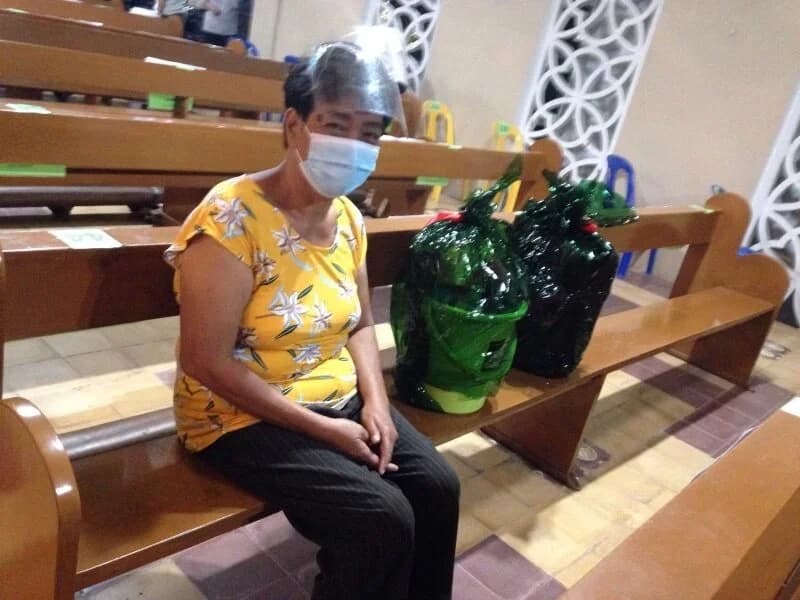 A woman in a face mask and face shield sits on a church pew with two wrapped Noche Buena baskets beside her.