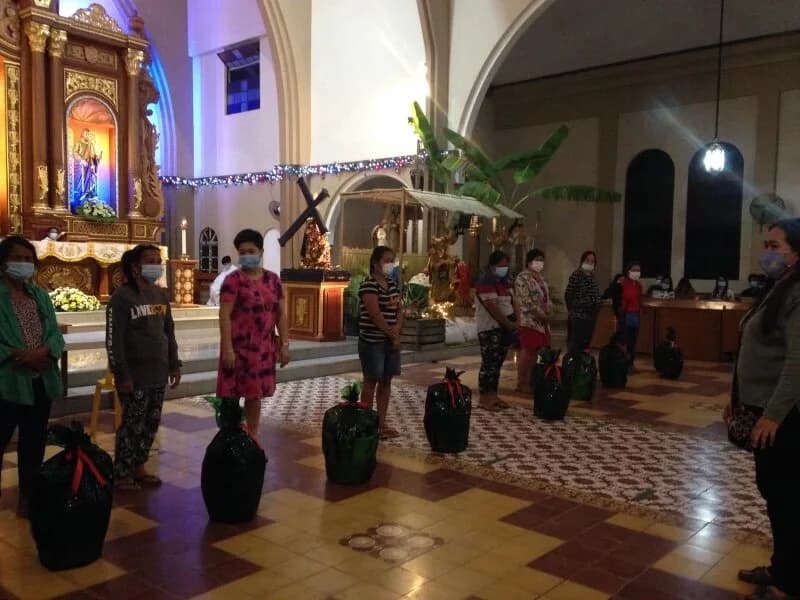 Women in face masks stand spaced apart in a church, a wrapped Noche Buena basket on the floor beside each of them.