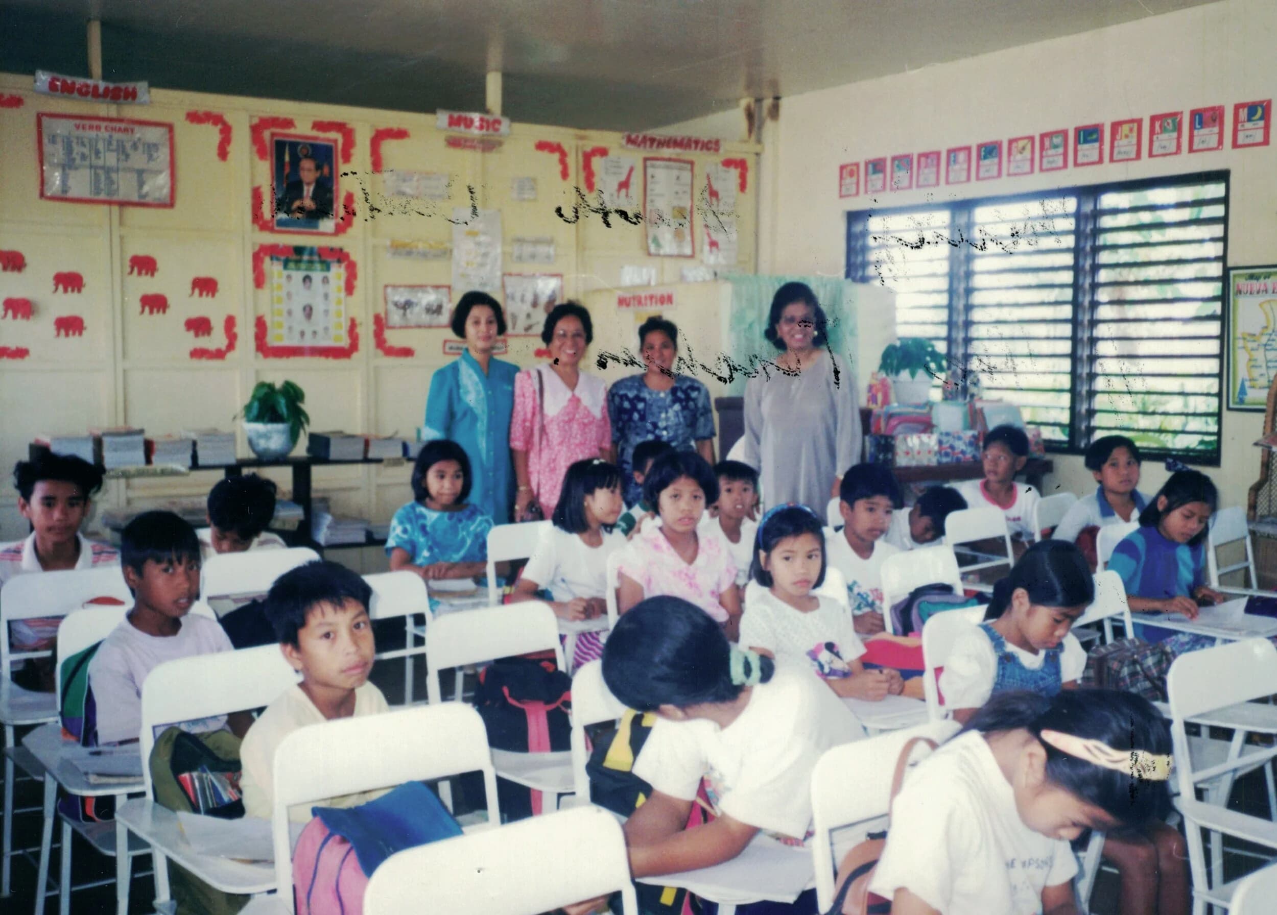 Scanned photo of a grade-school classroom: children seated at white desks, four women standing at the back beneath English, Music, and Mathematics posters.